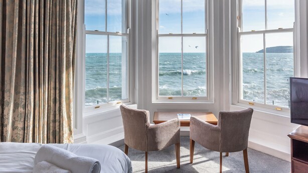 A room with a large window overlooking the sea, two chairs, a small table, and a bed. - The Queens Hotel (Penzance)