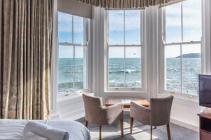 A room with a large window overlooking the sea, two chairs, a small table, and a bed. - The Queens Hotel (Penzance)