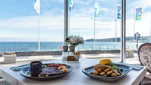 A table with plates of food and a vase of flowers, overlooking a scenic coastal view. - The Queens Hotel (Penzance)