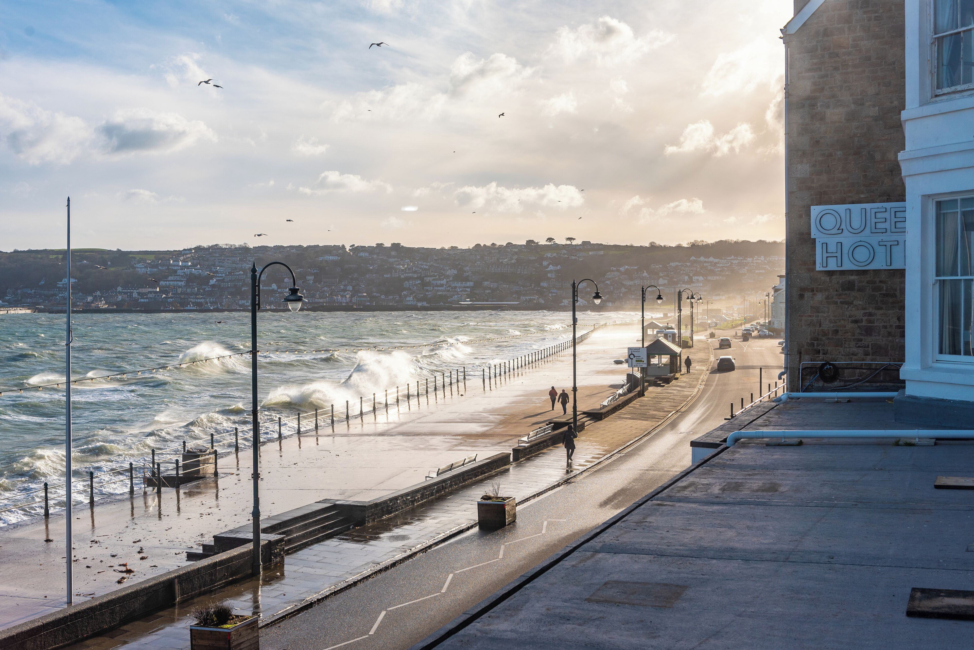 A coastal walkway with a view of the sea, a sign for 'Queen Hot', and a building with a stone facade.