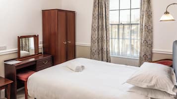 Quarto de hotel bem arrumado com cama, guarda-roupa de madeira, escrivaninha com espelho e janela com cortinas estampadas.