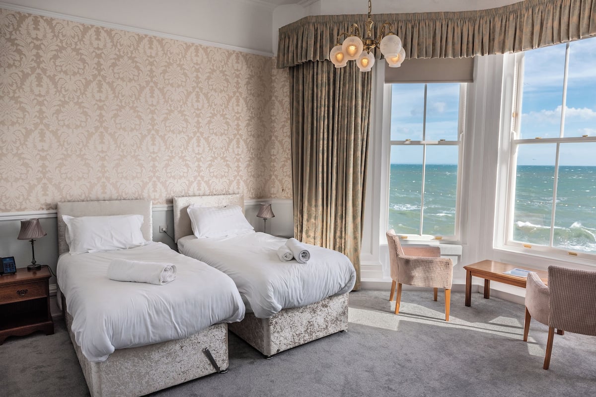 Habitación de hotel con dos camas, vista al mar, una araña y una pequeña mesa con una silla.