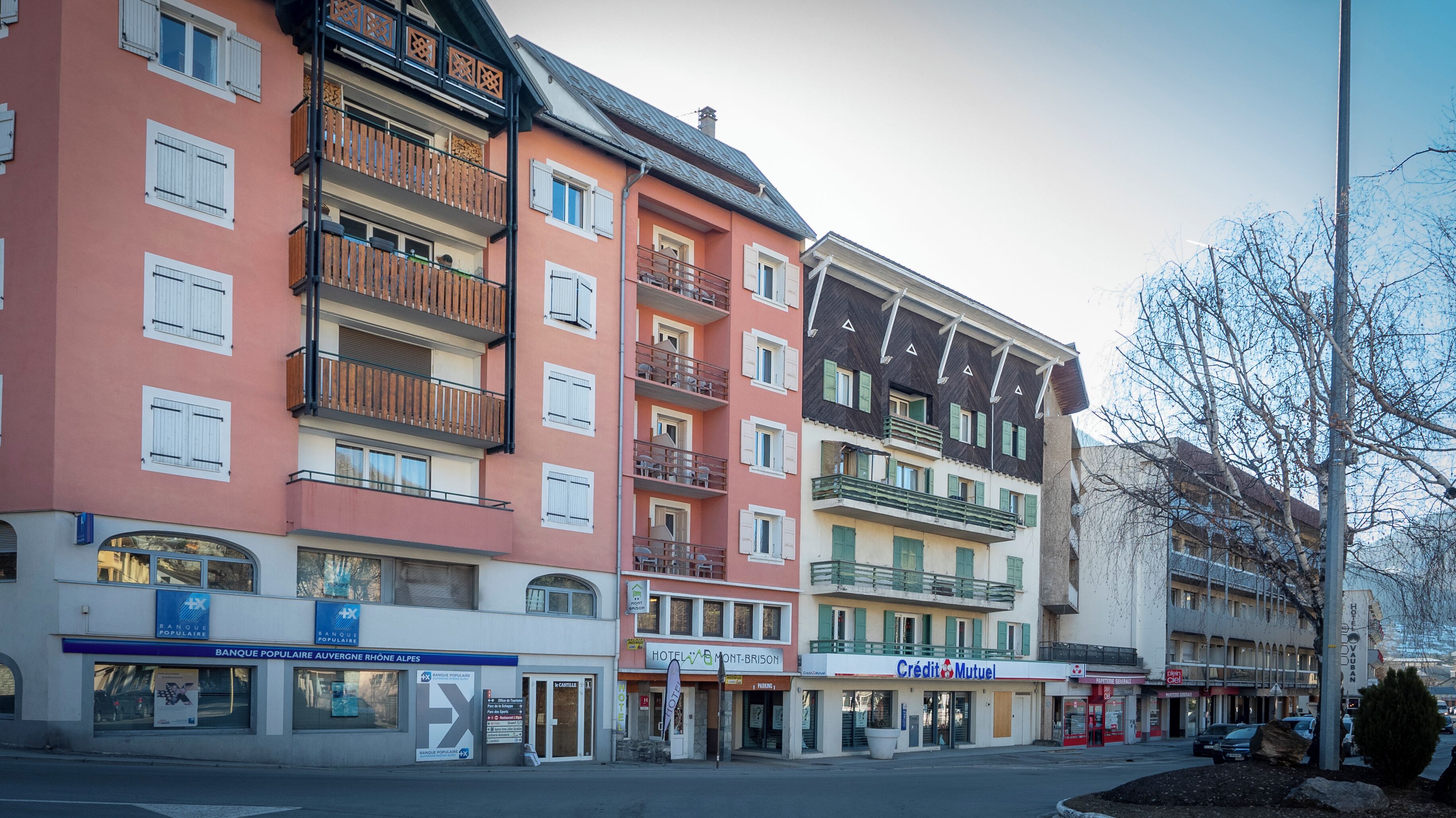 Un hotel de varios pisos con balcones, un restaurante a la vista y un banco en la planta baja.