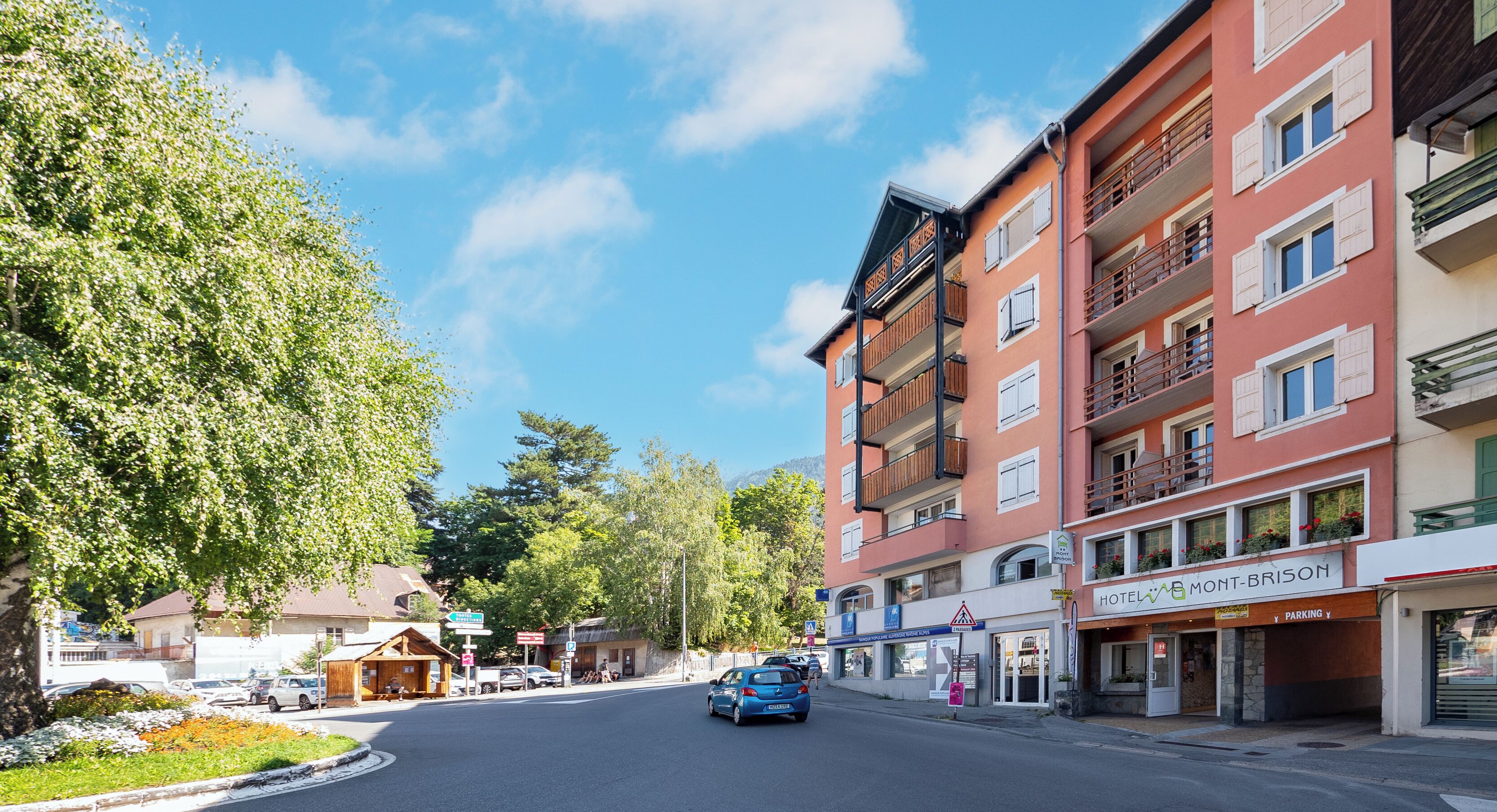 Une vue de rue avec une voiture bleue en mouvement, des immeubles dotés de balcons et un hôtel appelé « HÔTEL MONTR-BRISON ».