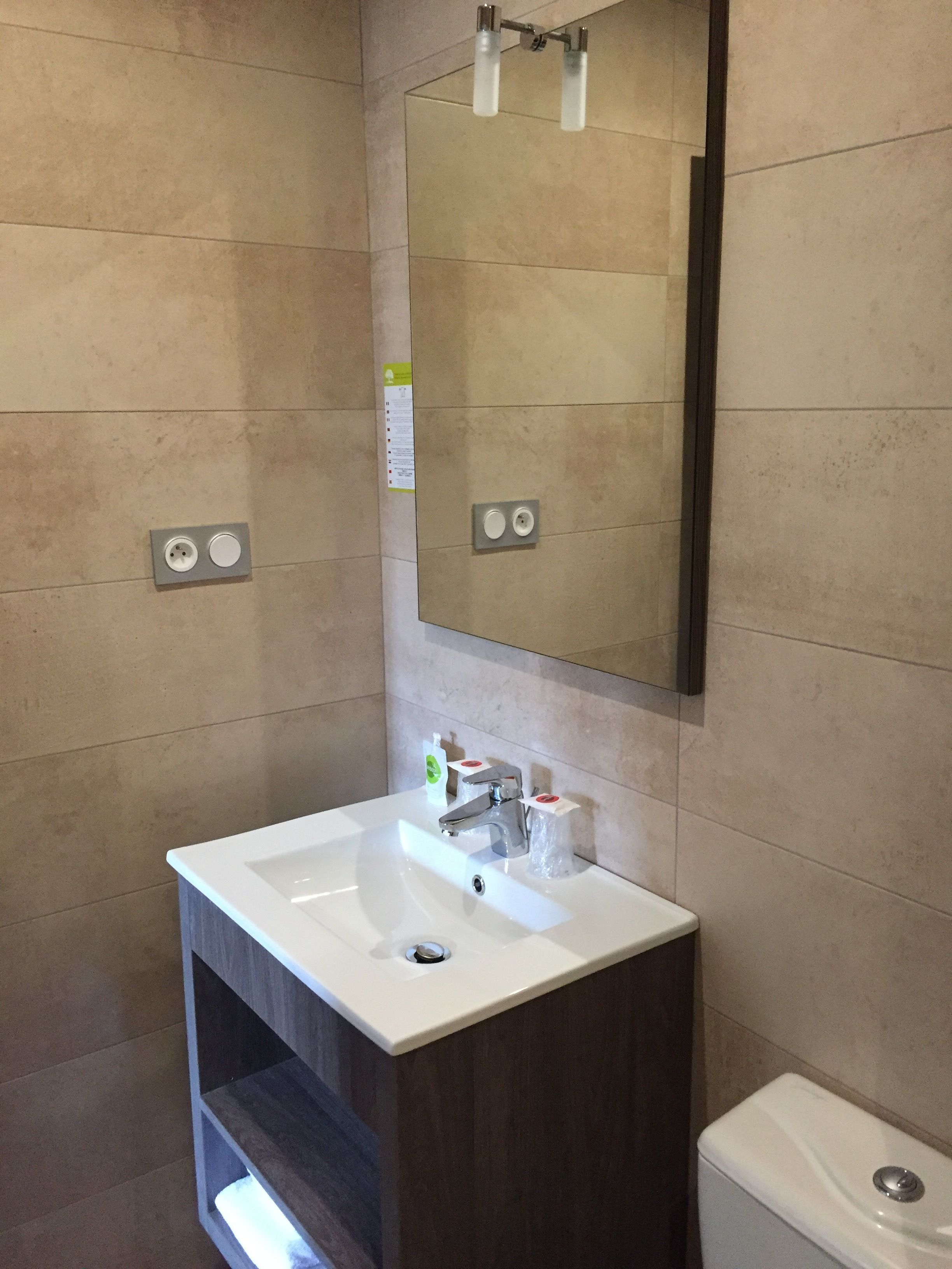 A modern bathroom with a white sink, a mirror, and a toilet. There are two electrical outlets on the wall.