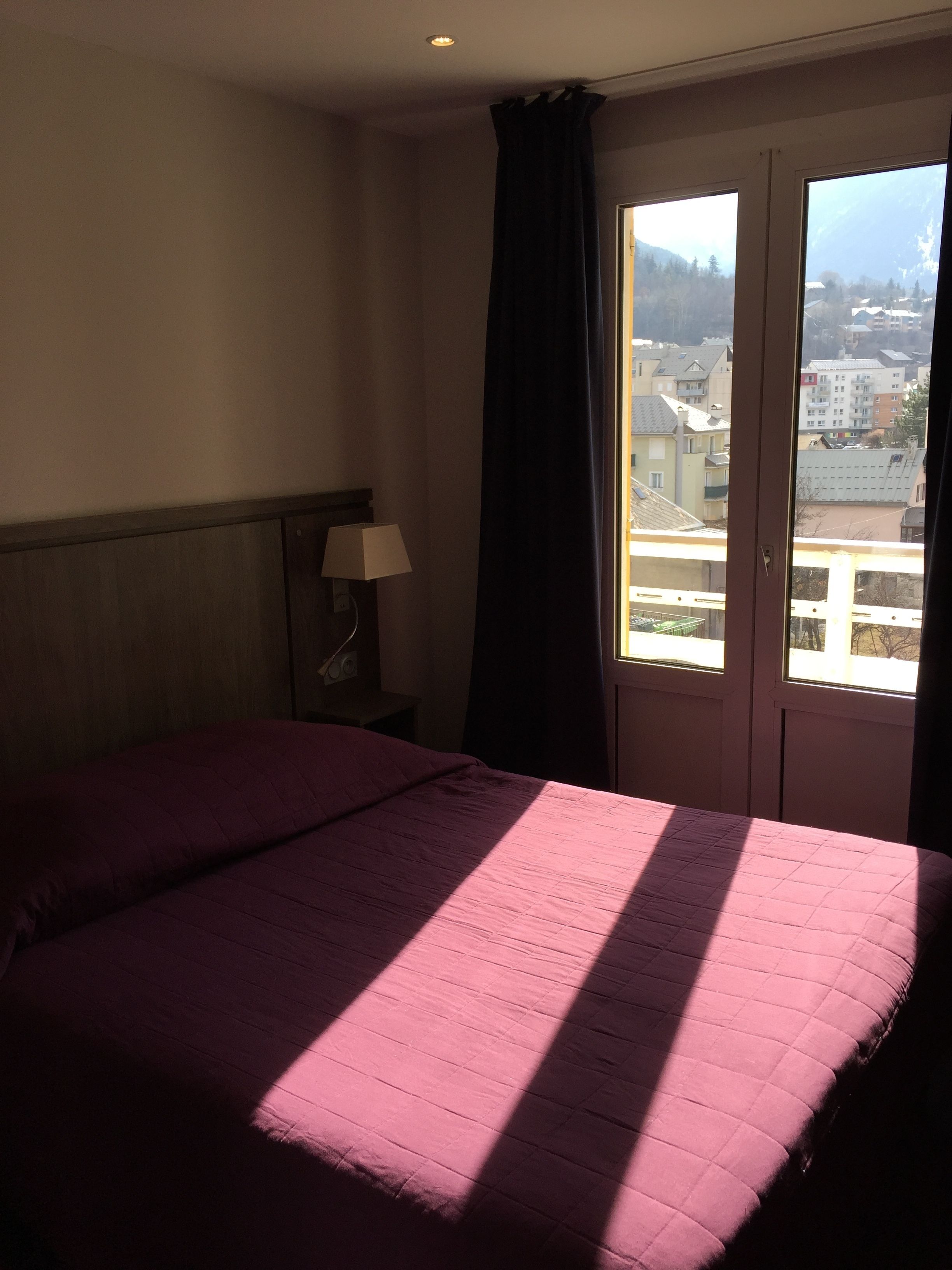 A hotel room with a bed, a nightstand, a lamp, and a window with a view of buildings.
