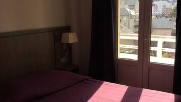 A hotel room with a bed, a nightstand, a lamp, and a window with a view of buildings.