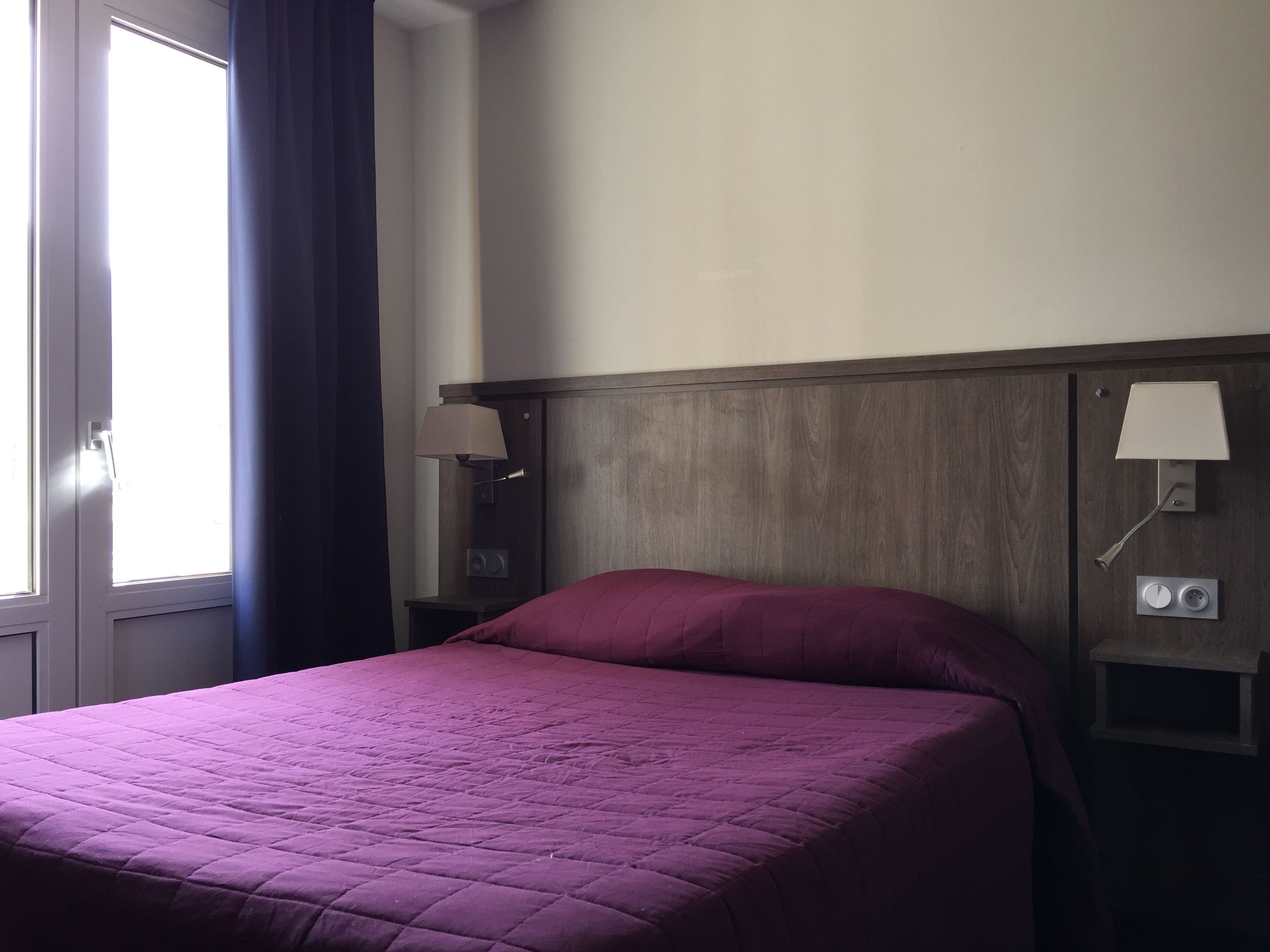 A hotel room with a bed, a nightstand with a lamp, and a wall-mounted light.