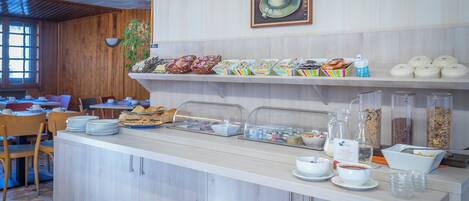 Un bufé de desayuno con diferentes alimentos, como pasteles, cereales y bebidas, en una habitación luminosa con detalles de madera y un ventanal grande.