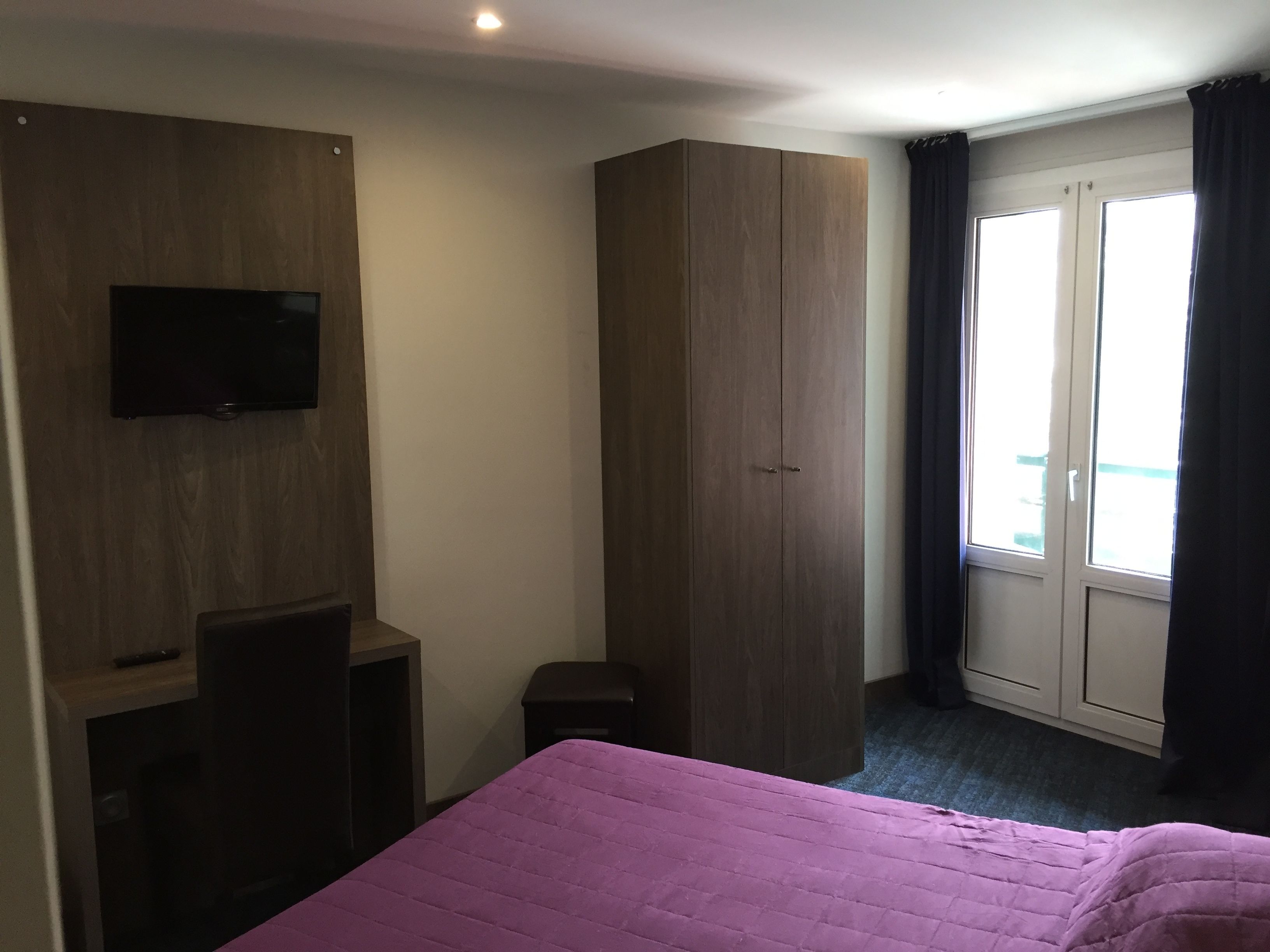 A hotel room with a bed, a wardrobe, a television mounted on the wall, and a window with curtains.