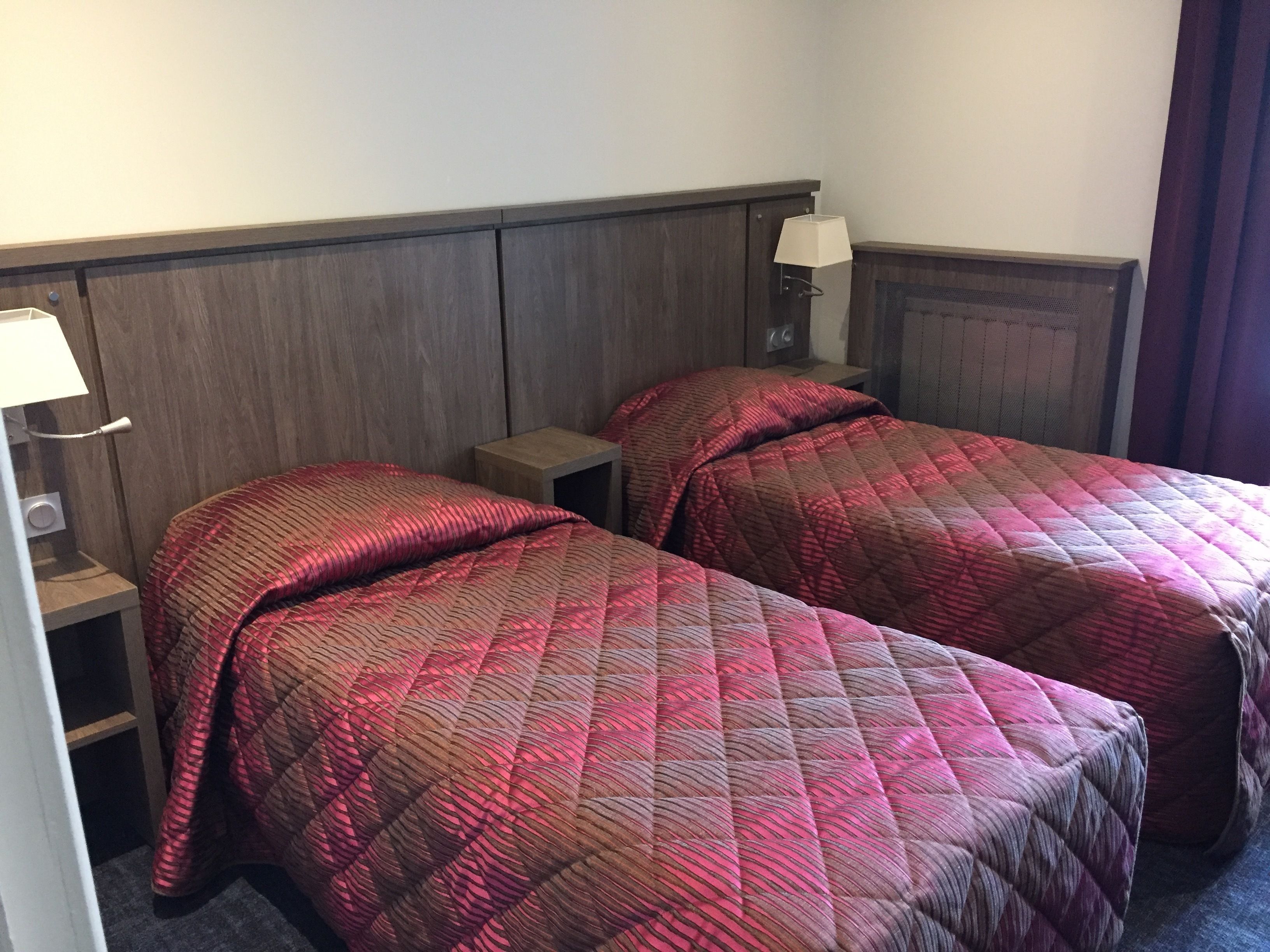A hotel room with two single beds, each with a red quilted bedspread and a nightstand with a lamp.