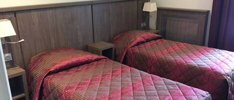 A hotel room with two single beds, each with a red quilted bedspread and a nightstand with a lamp.