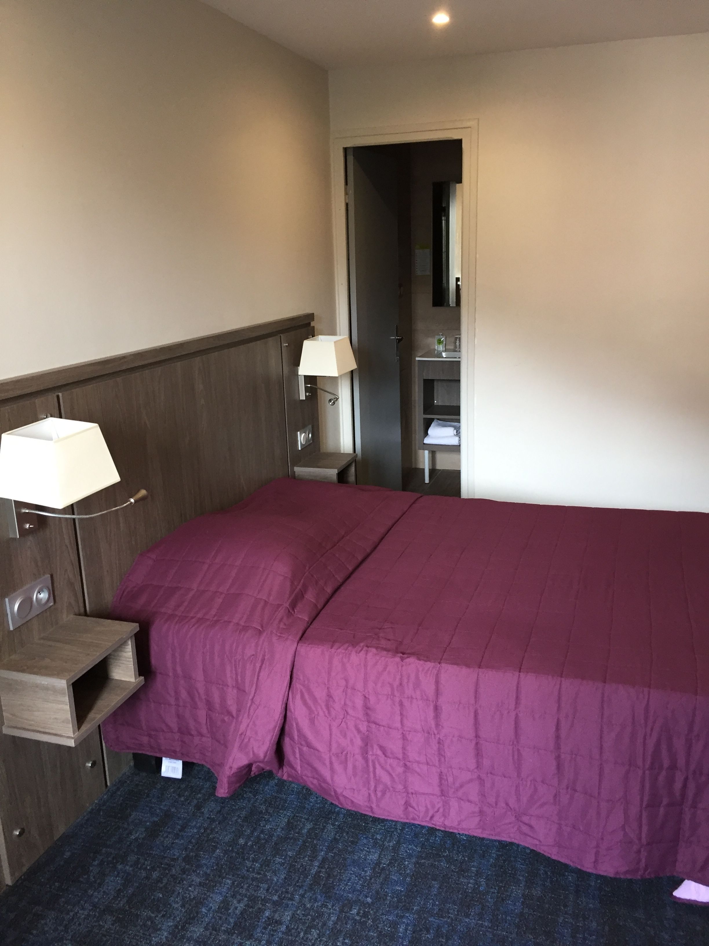Ein Hotelzimmer mit Bett, Nachttisch, Lampe, Spiegel und einem durchgegangene Tür sichtbaren Badezimmer.