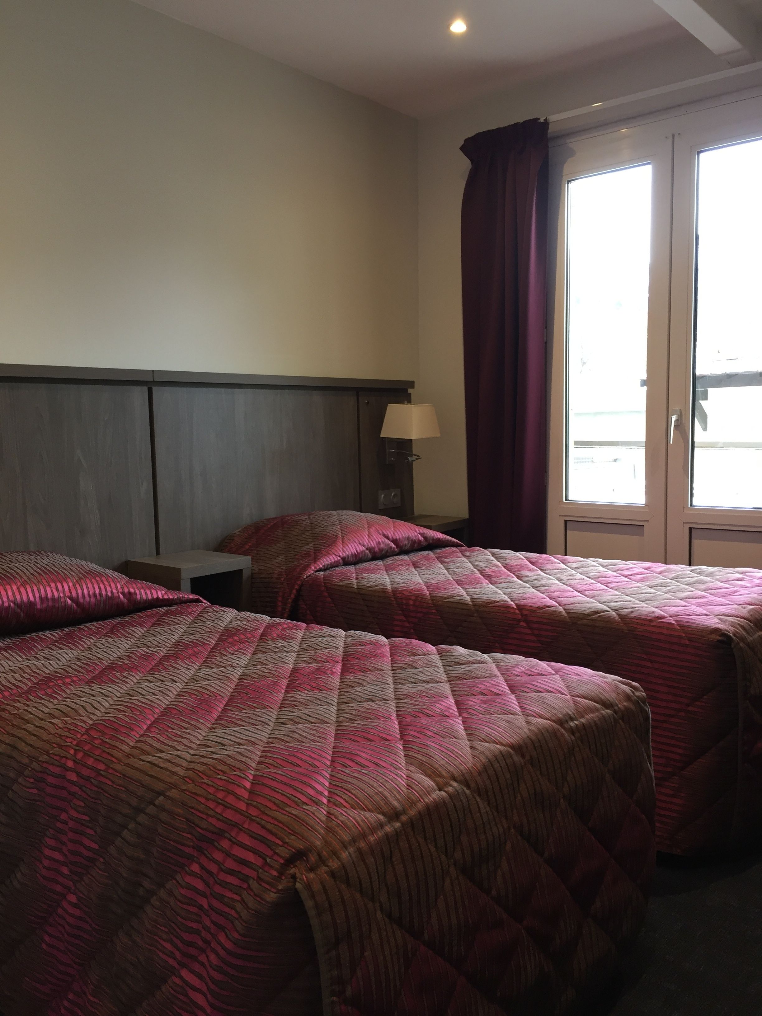A hotel room with two beds, a nightstand, a lamp, and a window with curtains.