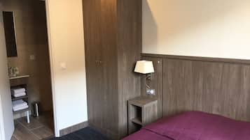 A hotel room with a bed, a wardrobe, a bedside table with a lamp, a small shelf with toiletries, and a trash bin.