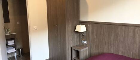 A hotel room with a bed, a wardrobe, a bedside table with a lamp, a small shelf with toiletries, and a trash bin.