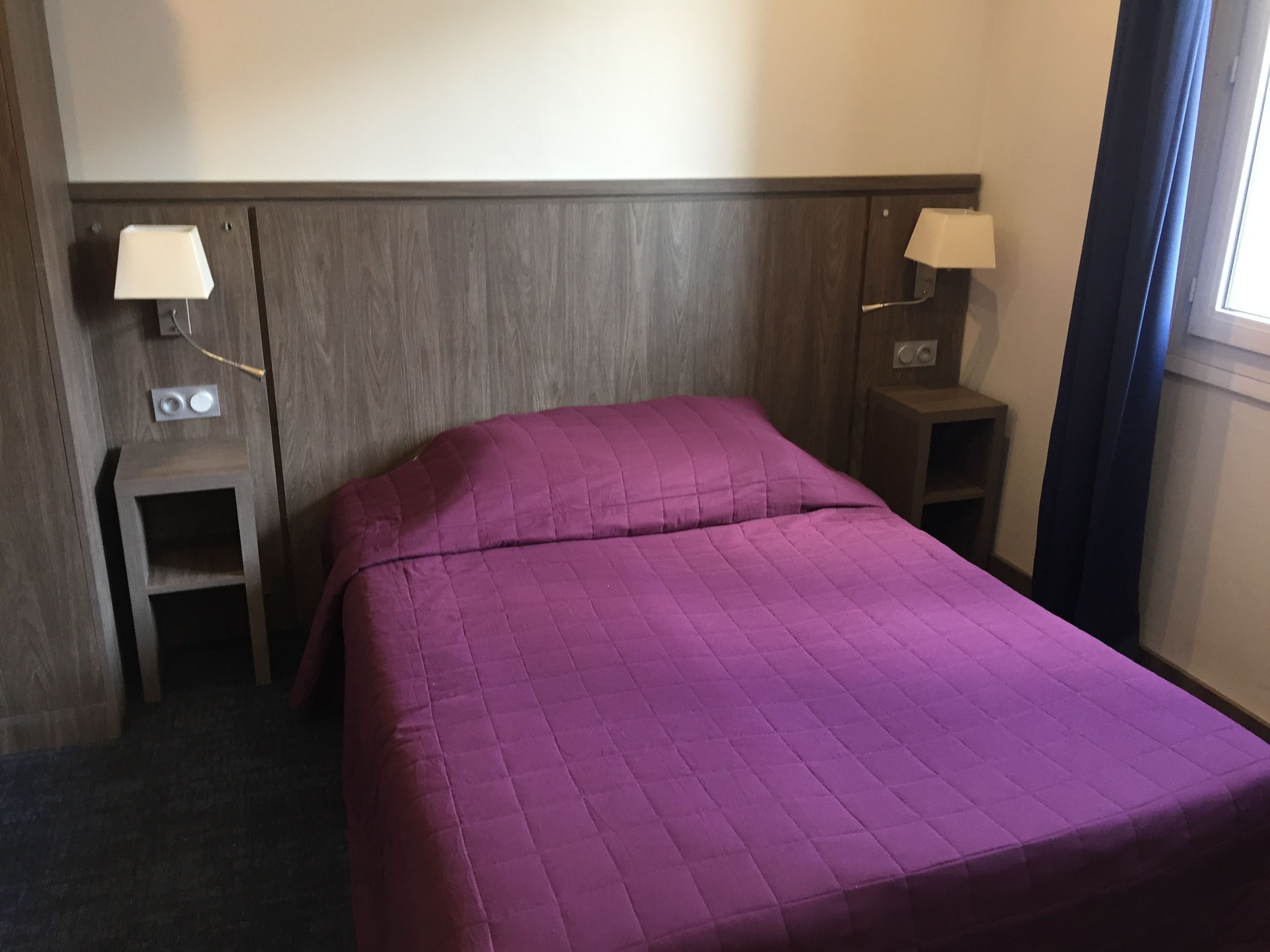 Ein Hotelzimmer mit einem Bett, zwei Nachttischen, einer Lampe auf jedem Tisch und einem Fenster mit Vorhängen.