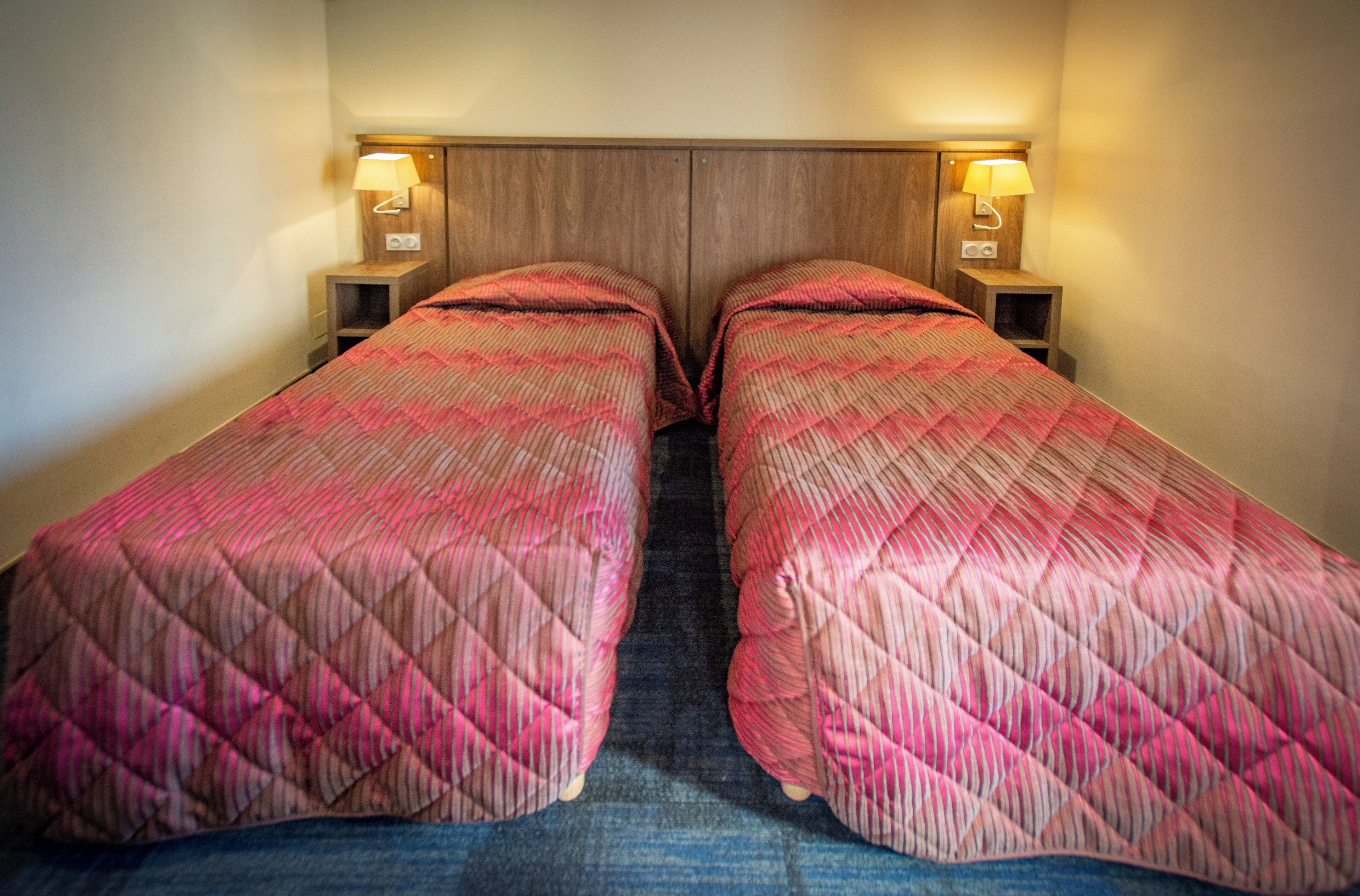 Habitación de hotel con dos camas individuales, cada una con cabecero de madera y lámparas de noche.