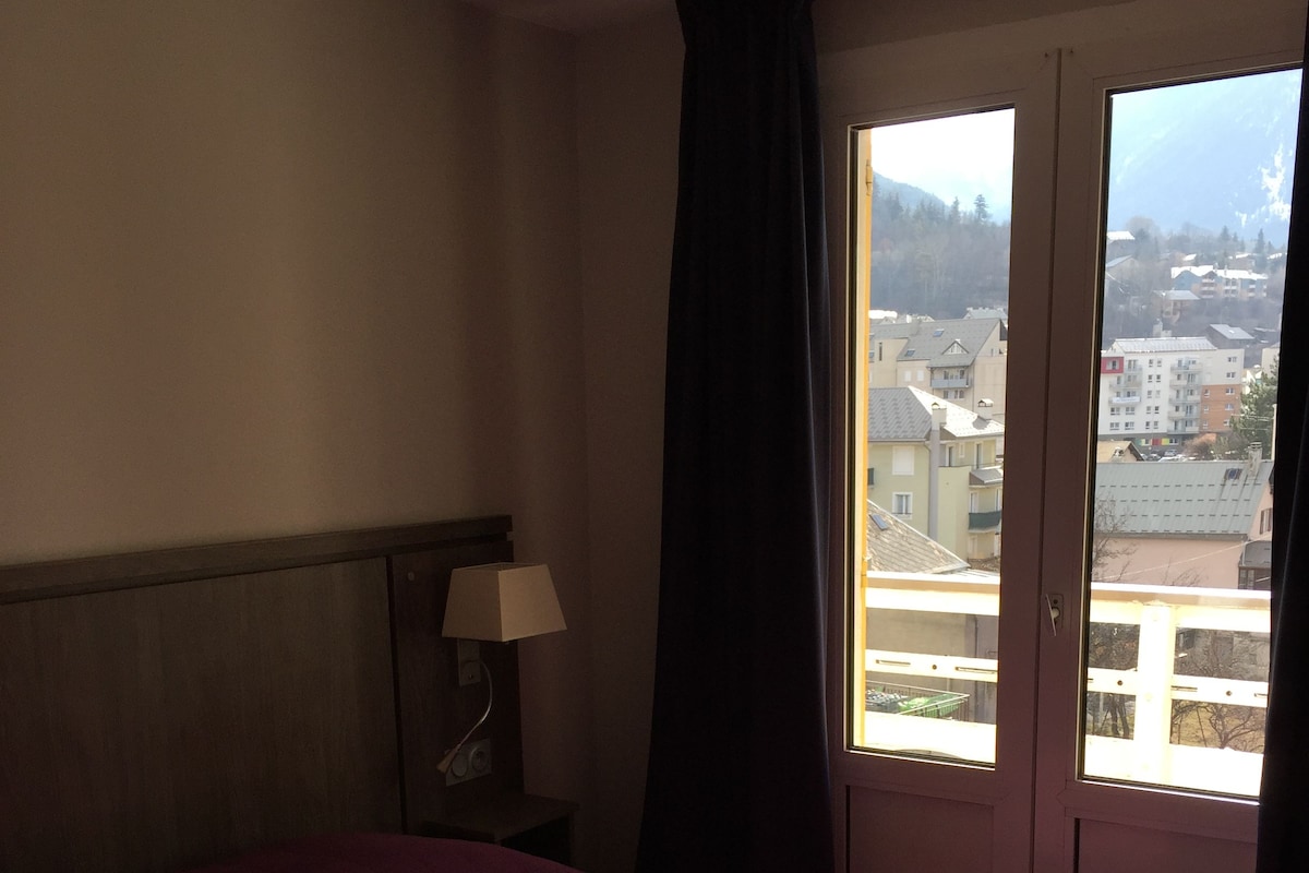 A hotel room with a bed, a nightstand with a lamp, and a window with a view of buildings and mountains.