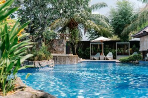A swimming pool with a waterfall feature, surrounded by lush greenery and palm trees. - The Leaf Jimbaran (Jimbaran)