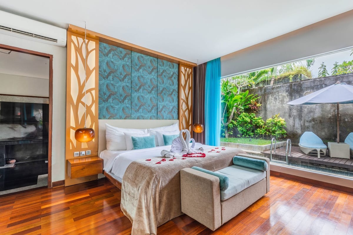 A hotel room with a bed, bedside tables, a sofa, a window with a view of greenery, and a balcony with seating.