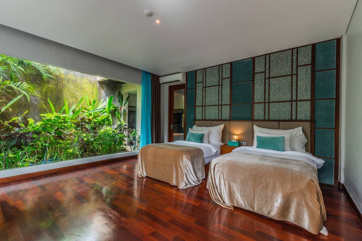 A hotel room with two beds, a large window with a view of greenery, and a traditional-style headboard.