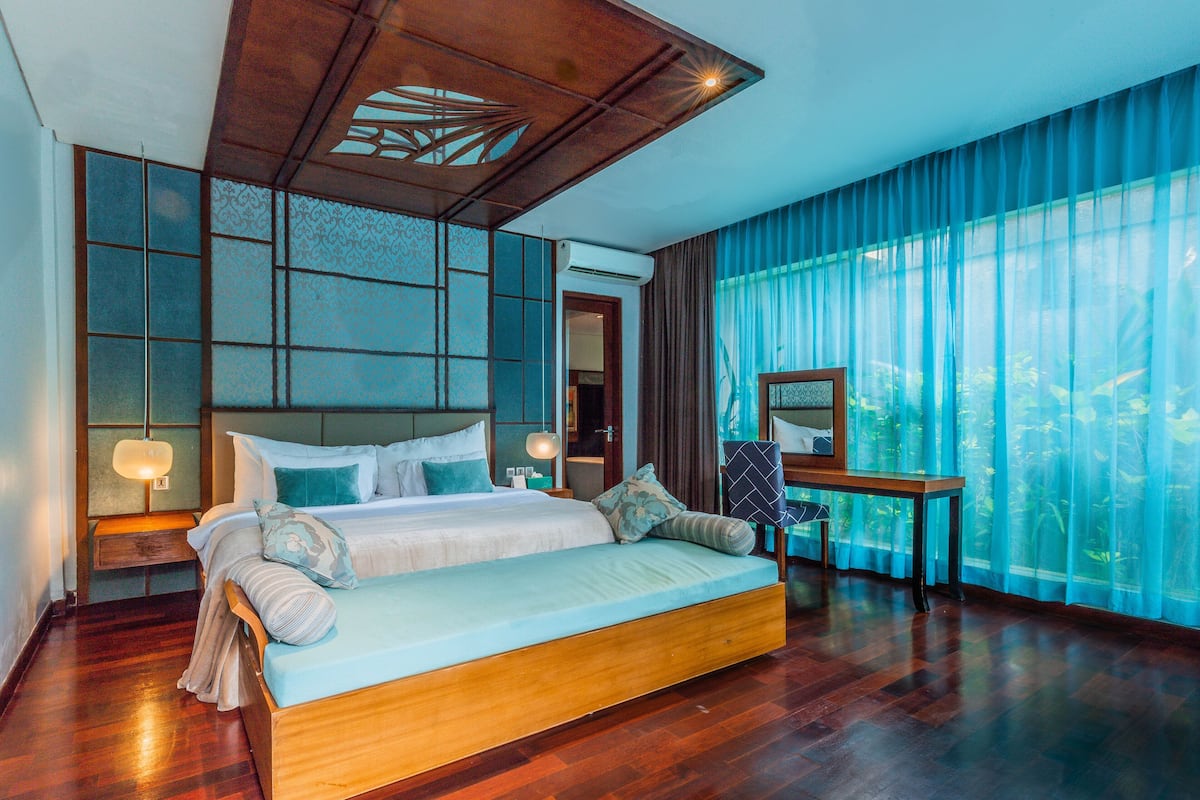 A spacious bedroom with a large bed, wooden headboard, and a view of greenery through sheer curtains.