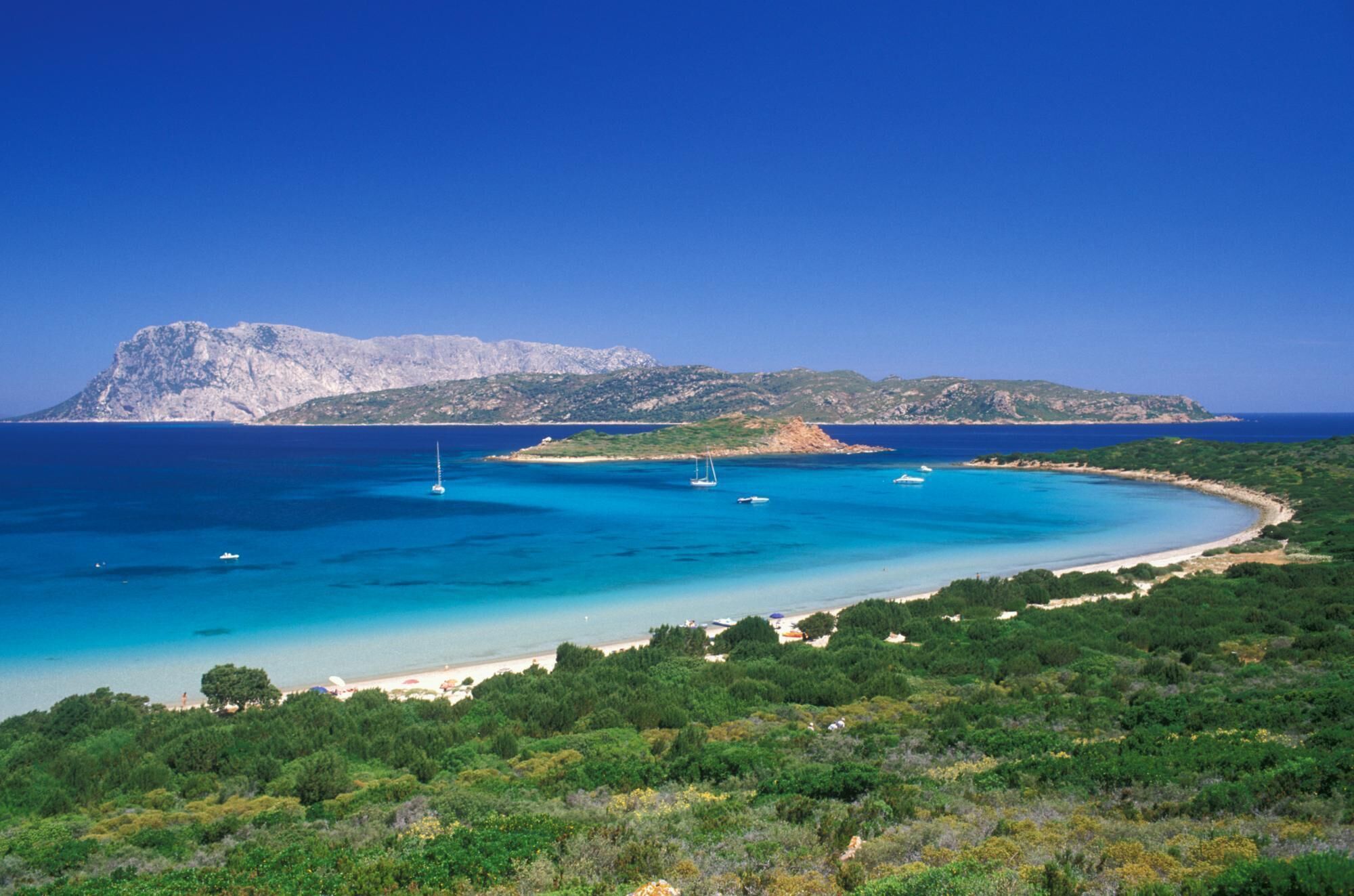 A coastal landscape with a clear blue sea, a sandy beach, and a mountainous backdrop.