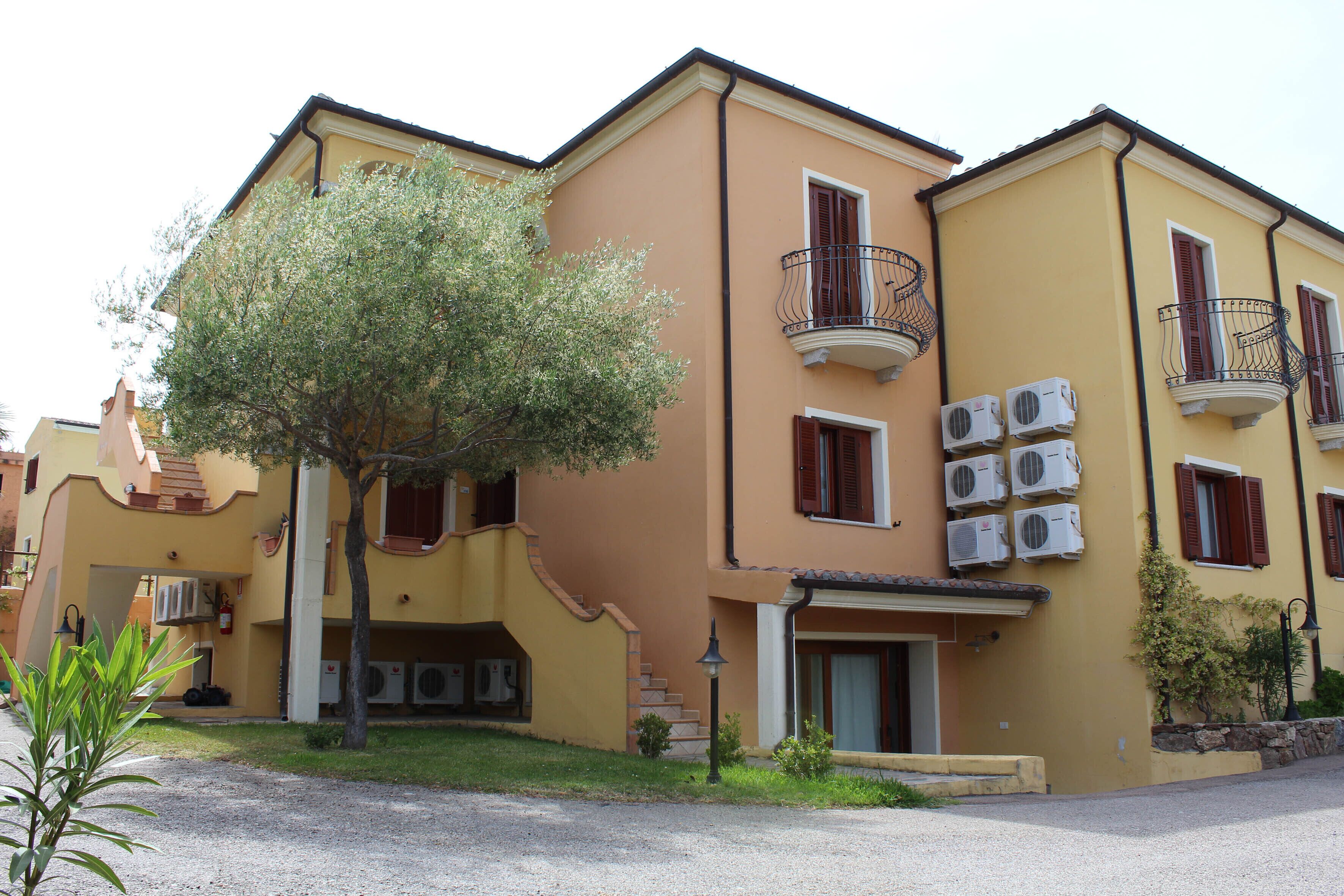 A yellow building with multiple balconies, air conditioning units, and a small tree in the front yard.