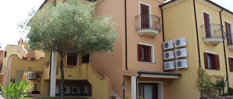 Un edificio amarillo con varios balcones, equipos de aire acondicionado y un pequeño árbol en el jardín delantero.