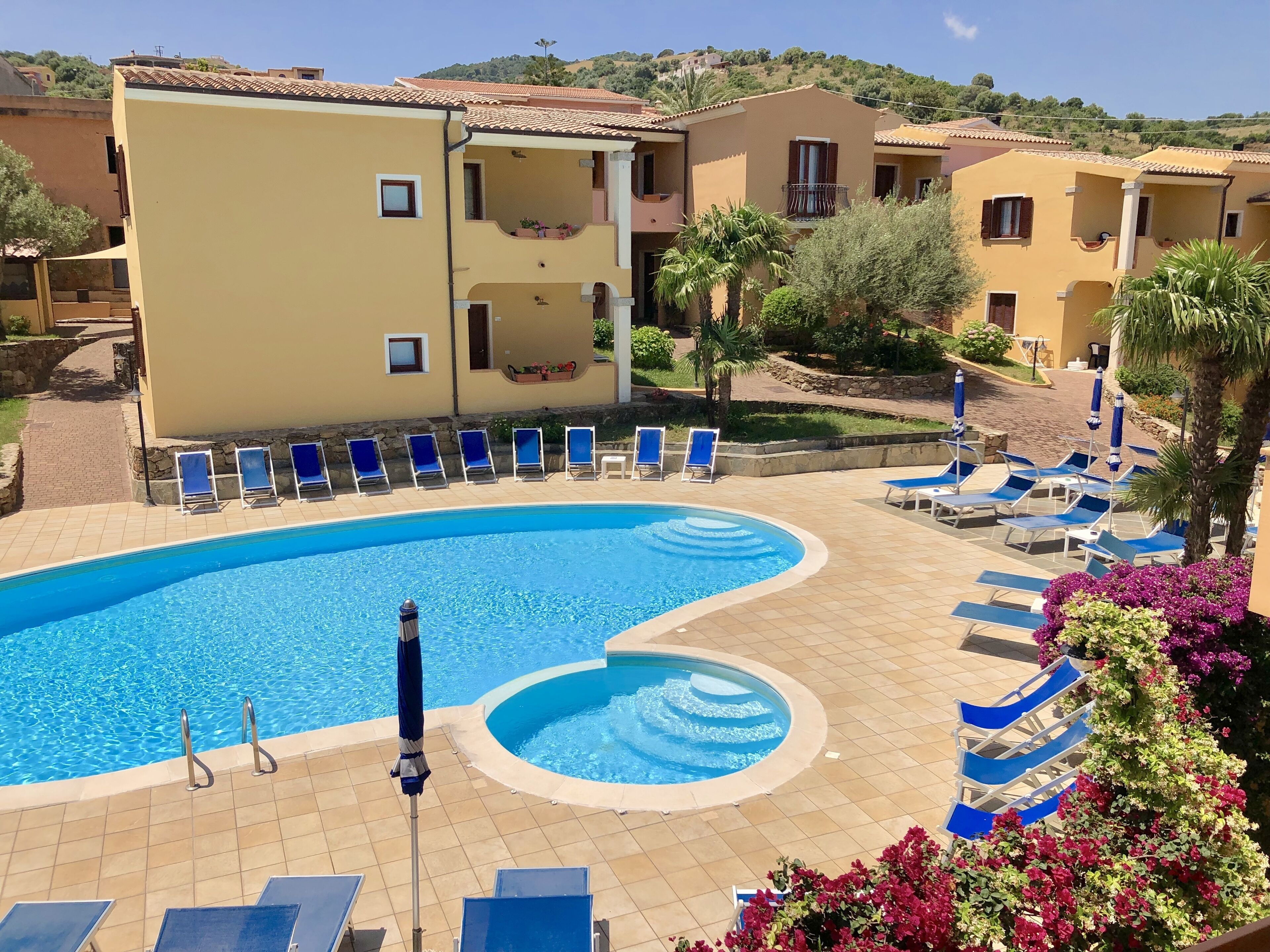 A swimming pool with blue lounge chairs and umbrellas, surrounded by buildings with balconies and a view of hills.