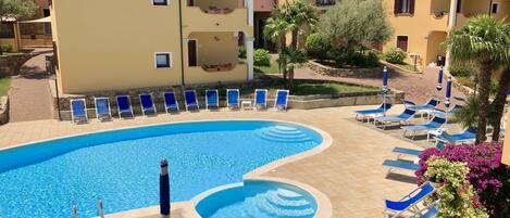A swimming pool with blue lounge chairs and umbrellas, surrounded by buildings with balconies and a view of hills.