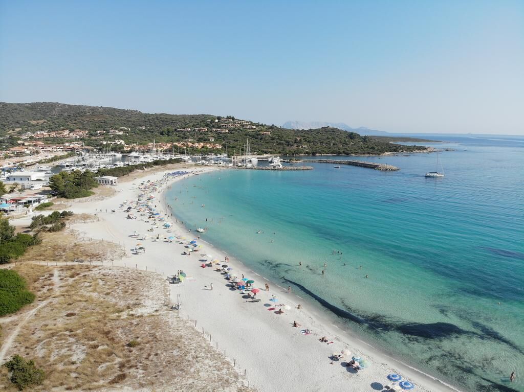 A coastal beach with clear turquoise waters, a sandy shore, and a marina with boats.