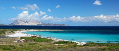 A coastal area with a beach, clear turquoise waters, and a mountain in the background.