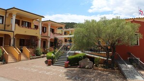 A multi-story building with a central staircase, outdoor seating area, and a flag. - Residence Corte dei Venti (Budoni)