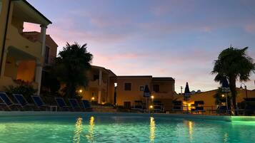 Una piscina con lettini e ombrelloni, circondata da edifici e palme.