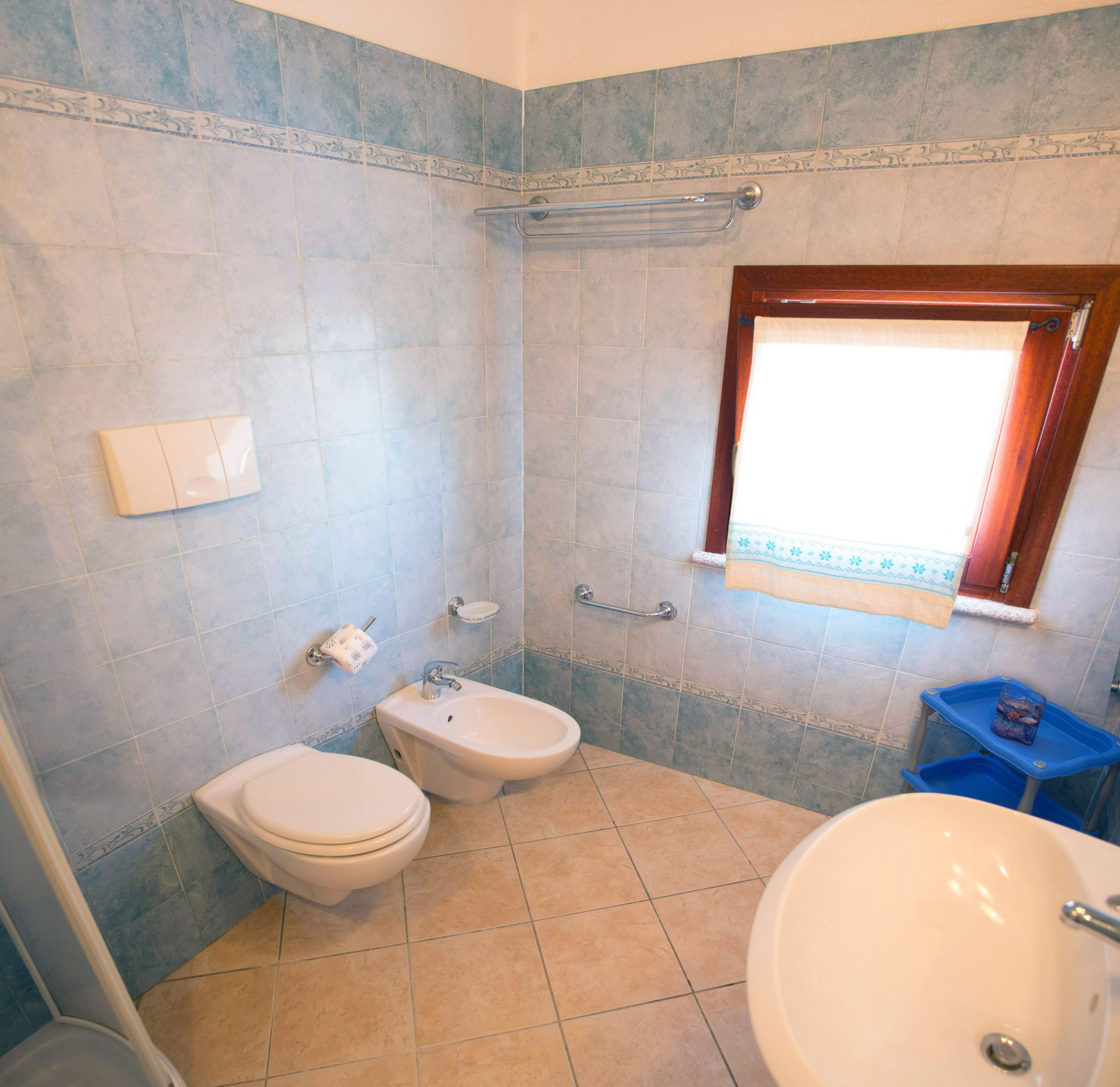 A bathroom with a toilet, bidet, sink, and a window with a curtain.