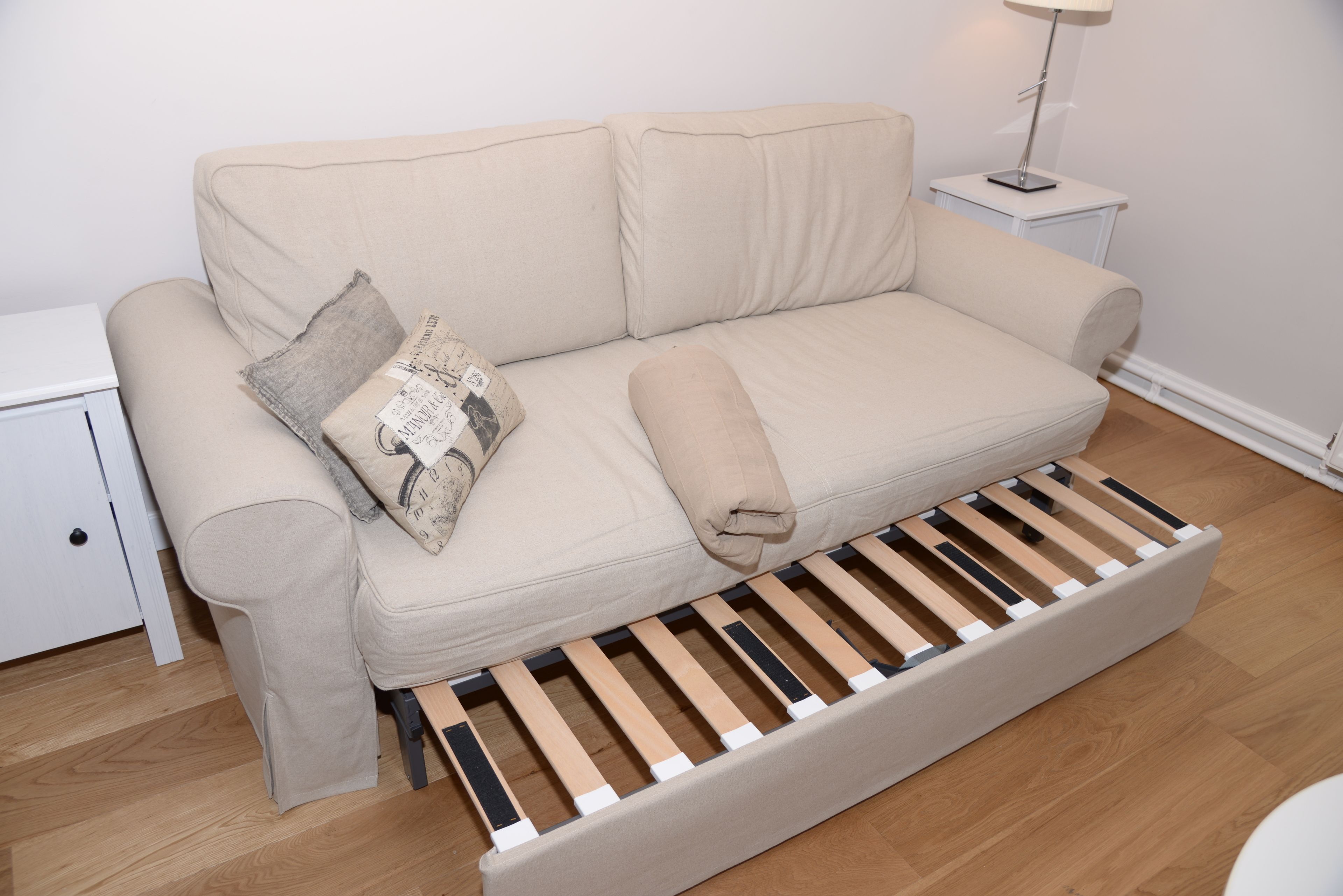A beige sofa with a wooden bench underneath, a small white nightstand with a lamp, and a white wall.