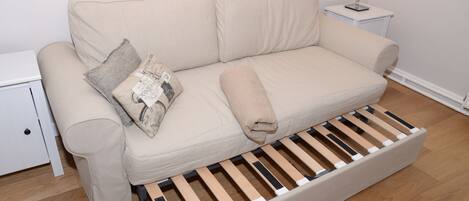 A beige sofa with a wooden bench underneath, a small white nightstand with a lamp, and a white wall.