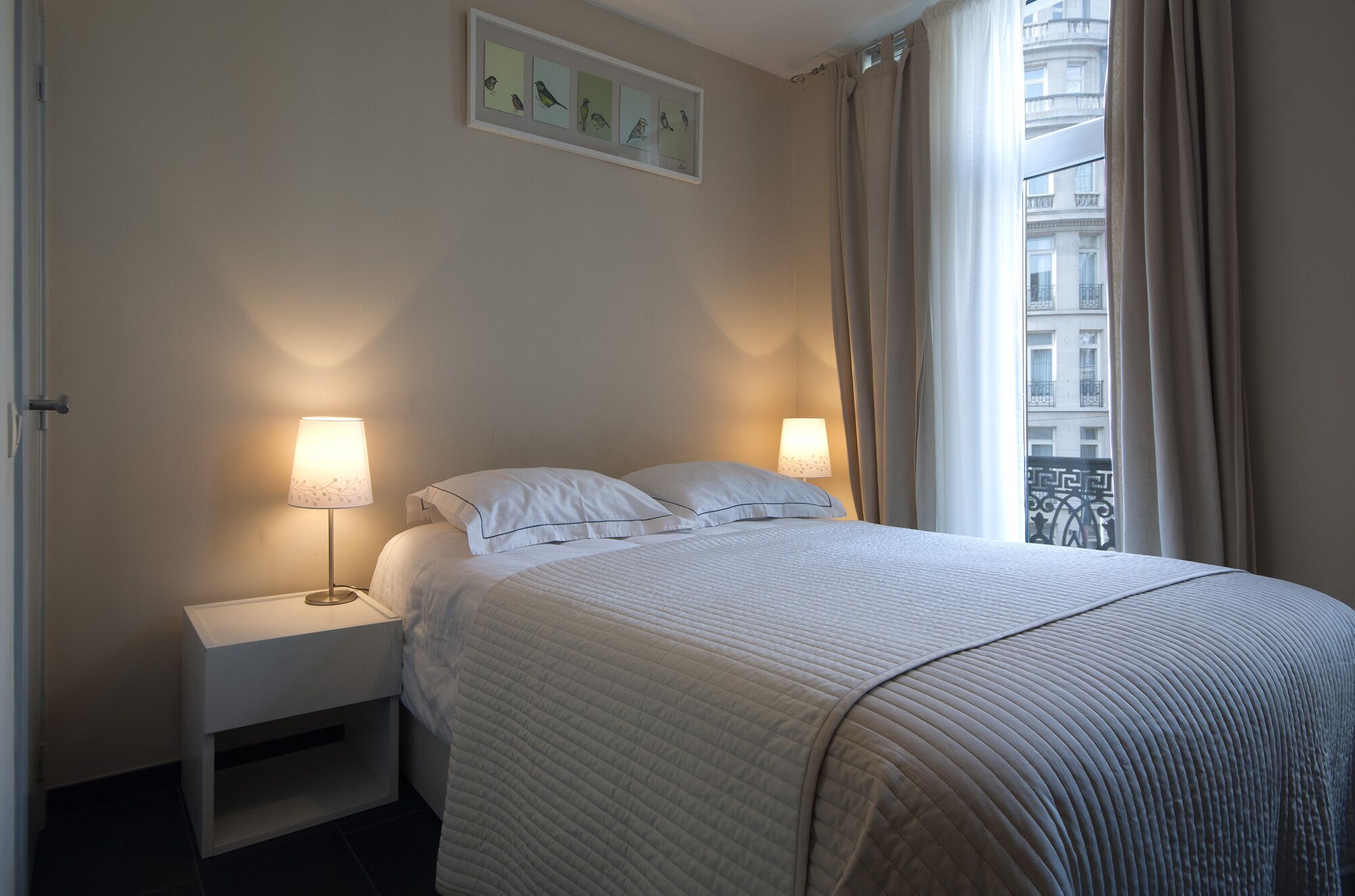 Een net opgemaakt bed met een gewatteerd deken, twee bedlampen, een klein nachtkastje en een lijst met een afbeelding van vogels aan de muur.