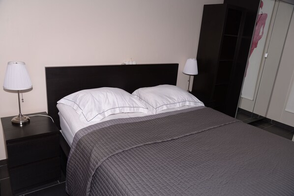 A neatly made bed with white linens and a gray quilted bedspread, a black nightstand with a lamp, and a wardrobe with a mirror.