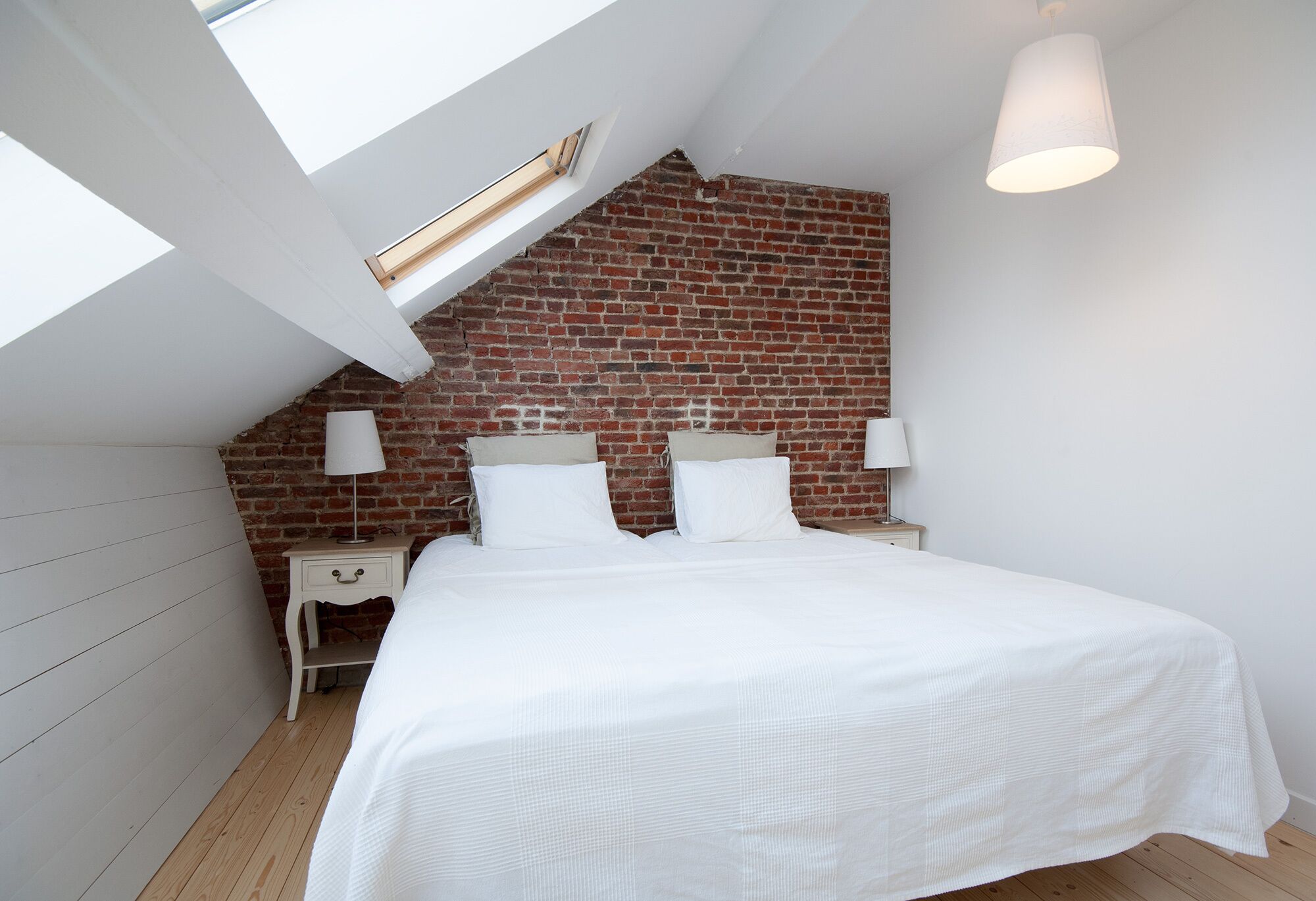 A bedroom with a sloped ceiling, exposed brick wall, a bed with white bedding, two bedside tables with lamps, and a skylight.