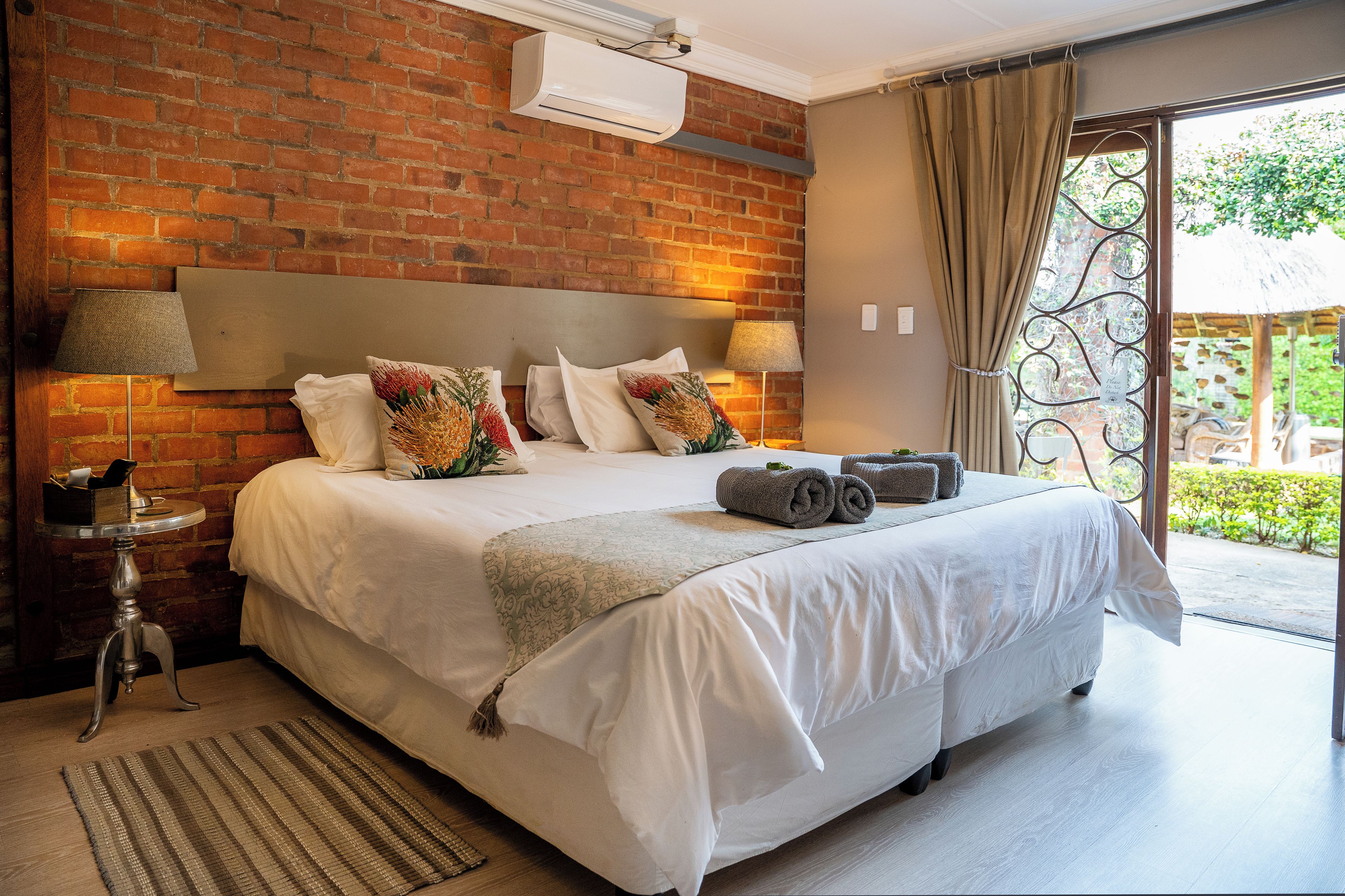 A bedroom with a brick wall, a large bed with white linens, two bedside lamps, and a view of the outdoors through a glass door.