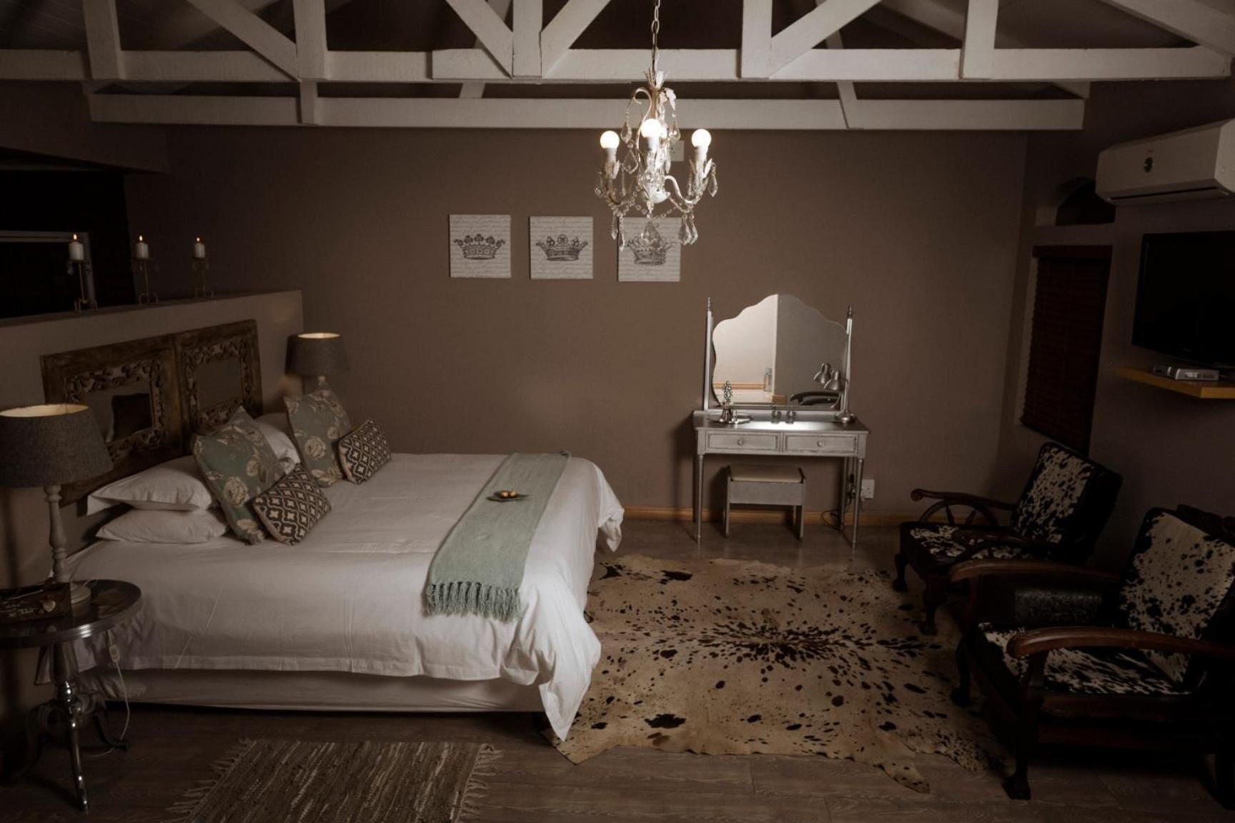 Une chambre à coucher avec un grand lit, une coiffeuse avec un miroir, un lustre et un téléviseur fixé au mur.
