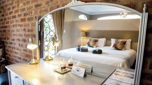 A hotel room with a large mirror, a bed with pillows, a bedside table with a lamp and a glass decanter, and a brick wall. - Klip en Kristal Guest House (Bela-Bela)