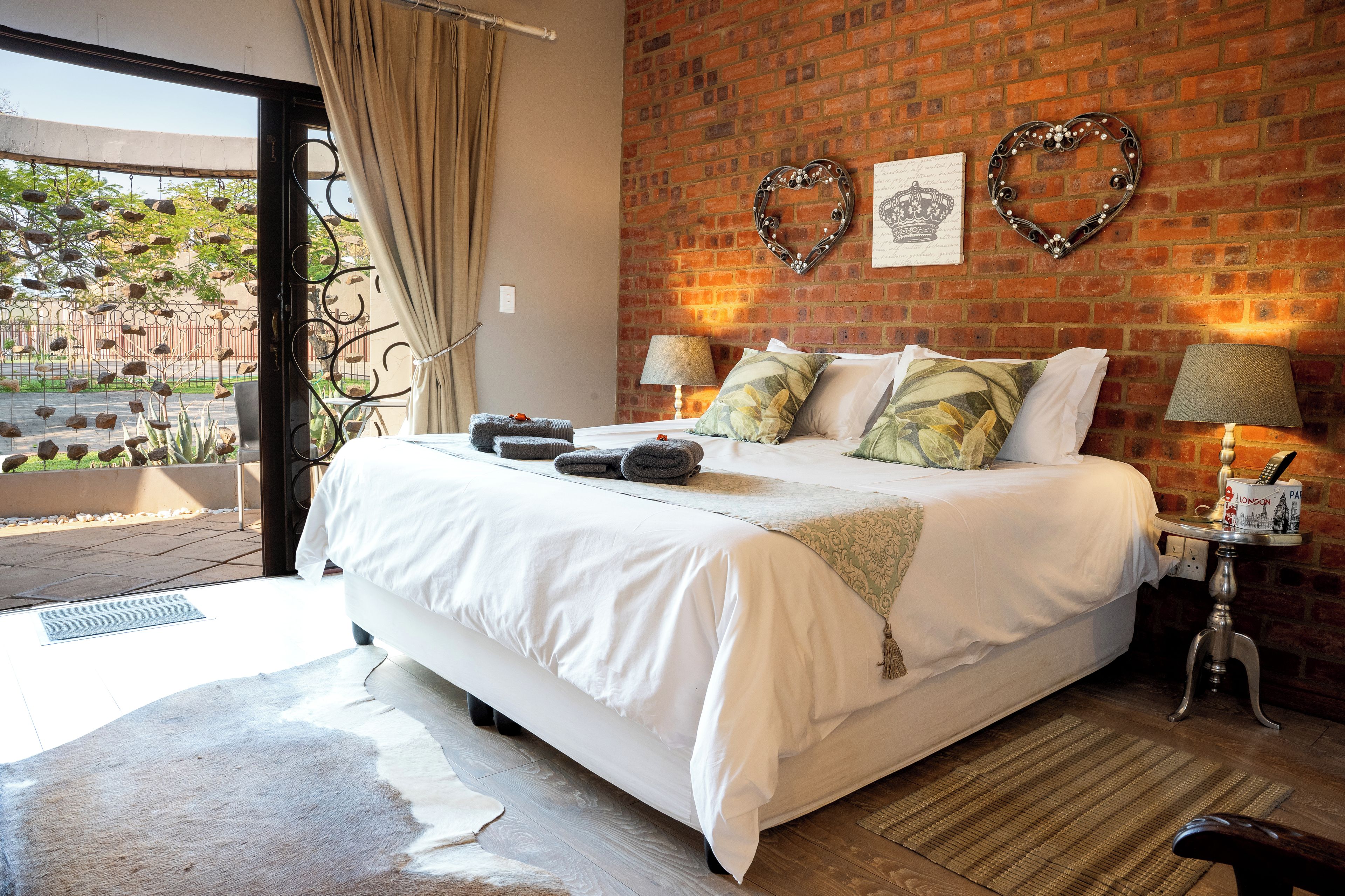 A bedroom with a large bed, a brick wall, two heart-shaped decorations, and a window with curtains.