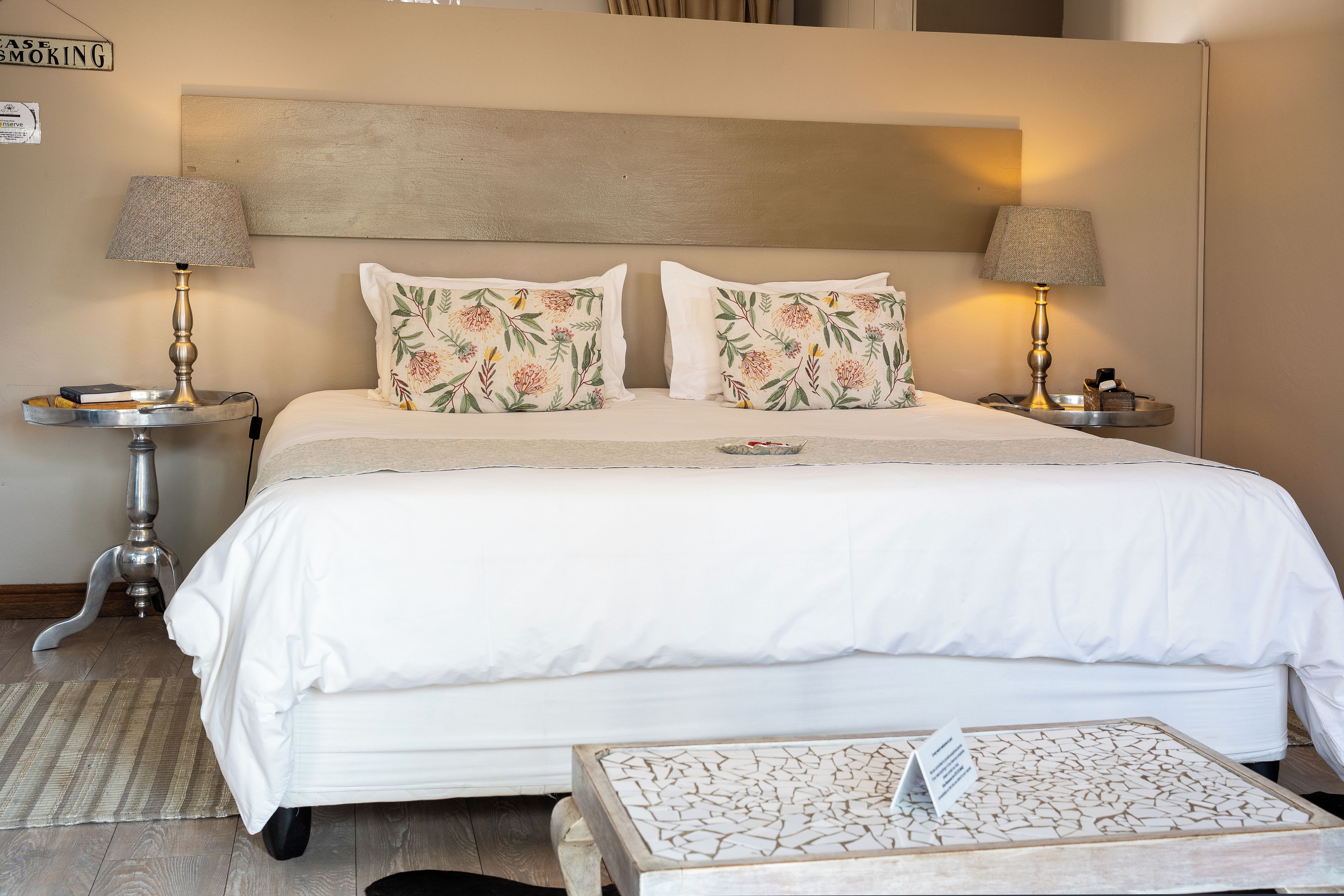 A neatly made bed with white bedding and floral pillows, two bedside lamps, a small table with a lamp, and a wall-mounted sign.