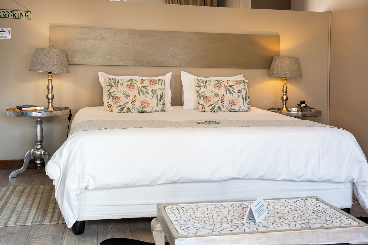 A neatly made bed with white bedding and floral pillows, two bedside lamps, a small table with a lamp, and a wall-mounted sign.