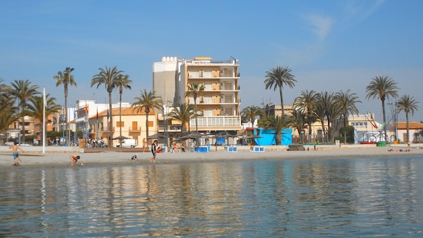 Eine Küstenlandschaft mit Strand, Palmen und einem mehrstöckigen Gebäude mit Balkonen.