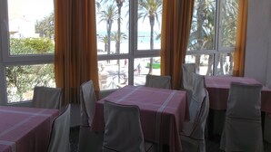 Ein Essbereich mit Tischen und Stühlen, großen Fenstern mit Vorhängen und Blick auf Palmen und einen Strand.